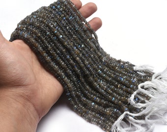 May include: Strands of gray labradorite gemstone beads with iridescent blue flashes. The beads are small, square-cut, and strung together. White tassels are visible at the end of the strands. The beads are held in a hand.