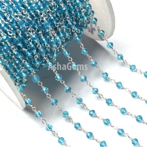 Puede incluir: Un carrete de cadena plateada con cuentas de cristal facetadas de color azul claro. La cadena está enhebrada con pequeñas cuentas redondas que están espaciadas uniformemente.