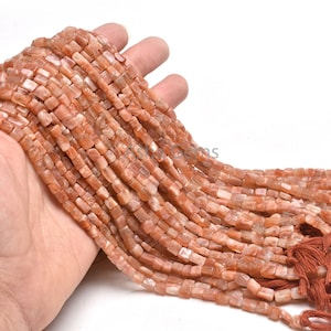 Puede incluir: Una hebra de cuentas de piedra de sol naranja, cada cuenta es de forma cuadrada con una superficie ligeramente irregular y facetada. Las cuentas están enhebradas en un cordón con un borla en el extremo.