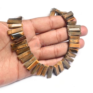 Puede incluir: Un collar de cuentas rectangulares de piedra preciosa ojo de tigre. Las cuentas son una mezcla de tonos marrón, dorado y gris, ensartadas juntas. Las piedras preciosas se sostienen en una mano sobre un fondo blanco.