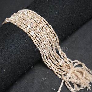 May include: Strands of small, round, beige beads with white highlights. The beads are strung together, forming multiple strands. The strands are draped over a black surface, with a tassel of beige thread at the end. The beads appear to be natural.