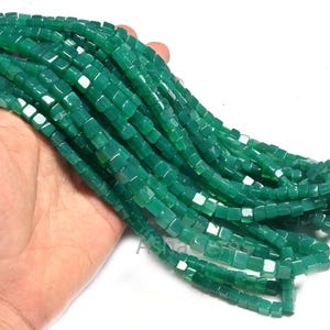 Puede incluir: Una hebra de cuentas de jade verde, cada cuenta tiene forma cuadrada. Las cuentas están enhebradas en un hilo transparente y se muestran en una mano.