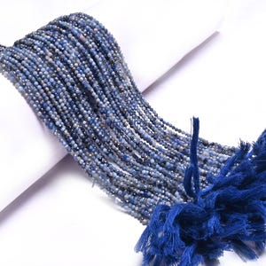 Könnte beinhalten: Stränge aus kleinen, facettierten blauen Perlen, mit einer tiefblauen Quaste am unteren Ende. Die Perlen haben eine Mischung aus blauen und grauen Farbtönen, die einen schimmernden Effekt erzeugen. Ideal für die Schmuckherstellung.