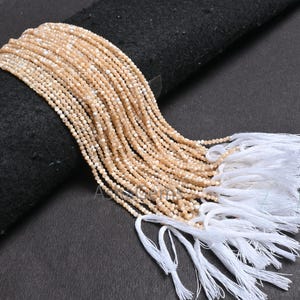 May include: A collection of small, light tan beads strung together, with white tassels at the end. The beads are draped over a black surface, creating a contrast in color and texture. The beads appear to be uniform in size and shape.