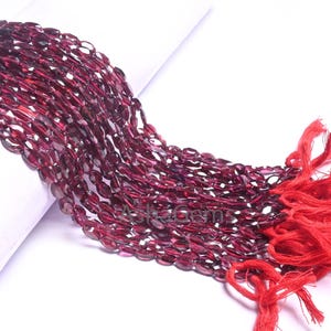 Può includere: Una collana di perle di granato rosso scuro, ogni perla è di forma ovale e sfaccettata. Le perle sono infilate su un filo rosso e hanno un fiocco all'estremità.