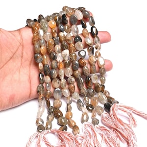 Può includere: Fili di perline di pietre preziose di forma ovale nei toni del marrone, beige e grigio. Le perline sono infilate insieme, con un nappa di filo bianco e rosso in basso. Le perline sono tenute in una mano su sfondo bianco.