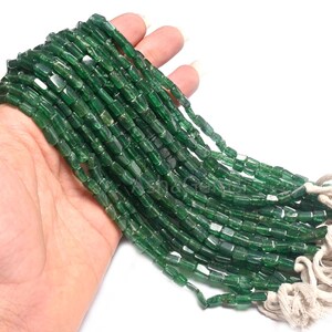 Puede incluir: Una hebra de cuentas de piedras preciosas verdes, cada cuenta tiene forma cuadrada con bordes ligeramente redondeados. Las cuentas están enhebradas en un cordón blanco con borlas en el extremo.