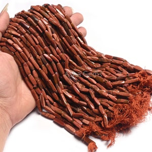 Könnte beinhalten: Eine Handvoll länglicher, rötlich-brauner Perlen mit einer glitzernden Textur. Die Perlen sind aufgefädelt, mit einer Quaste am Ende. Die Perlen haben eine warme, erdige Farbe, und der Gesamteindruck ist von natürlicher Schönheit. Die Perlen werden wahrscheinlich zur Schmuckherstellung verwendet.