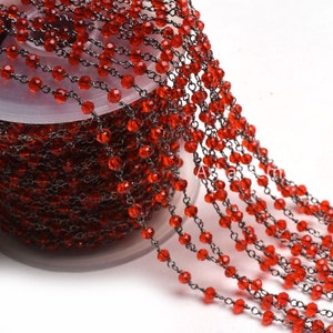 Puede incluir: Un carrete de cuentas de vidrio rojas enhebradas en una cadena plateada. Las cuentas son facetadas y tienen un acabado brillante.