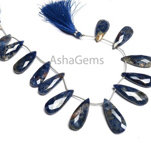 May include: Strands of faceted, teardrop-shaped blue gemstones with brown and white inclusions. The gemstones are strung together with silver-colored wire and are accented with blue tassels. The gemstones are arranged on a white background.