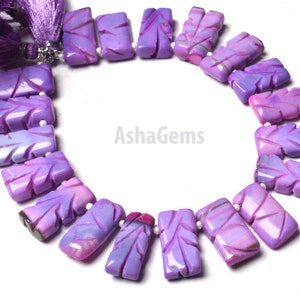 Puede incluir: Una hebra de cuentas de piedra preciosa talladas de color morado y rosa. Las cuentas son rectangulares y tienen un patrón de hojas talladas en ellas. Las cuentas están enhebradas en un cordón blanco y están atadas juntas con una borla morada.