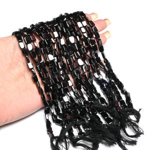 Peut inclure: Plusieurs brins de perles de pierres précieuses noires polies. Chaque perle est de forme rectangulaire aux bords arrondis. Les perles sont enfilées, formant plusieurs brins. Des pompons noirs maintiennent les brins. Fond blanc.