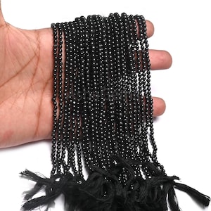 Puede incluir: Múltiples hebras de pequeñas cuentas redondas negras con un acabado brillante. Las cuentas están ensartadas y terminadas con borlas negras. Estas cuentas son probablemente para la fabricación de joyas o artesanía.