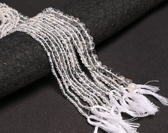 May include: A collection of clear quartz crystal bead strands with white tassels. The strands are arranged in a fan shape on a black surface.