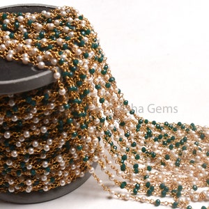 Peut inclure: Une bobine de chaîne perlée avec des perles blanches alternant avec des perles facettées vert foncé, enfilées sur une chaîne dorée. La chaîne est partiellement déroulée, révélant le design complexe. Les perles sont petites et espacées uniformément.