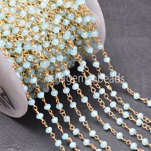 Puede incluir: Primer plano de una cadena de cuentas. Pequeñas cuentas facetadas de color azul claro ensartadas en alambre dorado. Las cuentas están espaciadas uniformemente, creando un aspecto delicado. La cadena se utiliza probablemente para la fabricación de joyas.