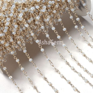 Puede incluir: Una hebra de cadena dorada con cuentas de ópalo blanco. La cadena está enrollada alrededor de un carrete y las cuentas cuelgan en cascada.