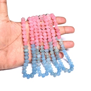 Peut inclure: Plusieurs brins de perles de pierres précieuses, allant du rose au bleu. Les petites perles rondes sont sculptées de motifs floraux. Les perles sont tenues dans une main sur fond blanc. Idéal pour la création de bijoux.