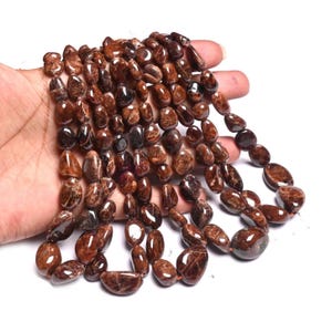 Könnte beinhalten: Stränge polierter, oval geformter, dunkelbrauner Edelsteinperlen. Die Perlen variieren in der Größe und haben eine glänzende Oberfläche. Die Edelsteine sind zu einer Halsketten- oder Armbandkomponente aufgefädelt. Die Perlen werden in einer Hand vor weißem Hintergrund gehalten.