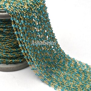 May include: A spool of teal blue faceted glass beads strung on a gold chain. The chain is made of small, delicate links. The beads are uniform in size and shape. The chain is wound around a gray spool.