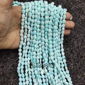 Puede incluir: Una hebra de cuentas de piedras preciosas de color azul claro, cada cuenta es lisa y de forma ovalada. Las cuentas están enhebradas en un hilo transparente y tienen un diámetro aproximado de 6 mm.