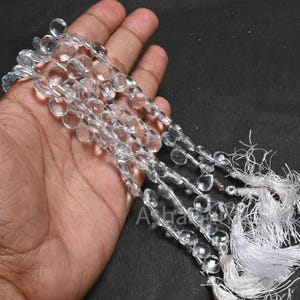 Könnte beinhalten: Eine Strähne aus klaren, facettierten und runden Quarzkristallperlen mit einem weißen Quasten am Ende.