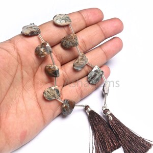 Puede incluir: Primer plano de cuentas de piedras preciosas en tonos grises, marrones y crema, con forma irregular. Las cuentas están ensartadas en un alambre plateado y terminadas con borlas marrones. Las cuentas se sostienen en una mano.
