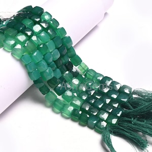Pode incluir: Um fio de contas de pedra de ágata verde, cada conta é facetada e de forma quadrada. As contas são enfiadas em um cordão com uma borla verde na ponta.
