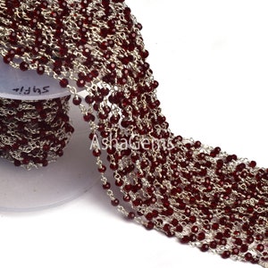May include: A close-up of a long strand of red gemstone beads strung on a silver chain. The beads are faceted and have a shiny finish. The chain is delicate and has a fine link pattern. The strand is coiled on a white spool with the word "Supply" printed on it.