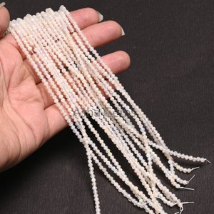 Peut inclure: Plusieurs brins de petites perles rondes d'opale blanche. Les perles ont un léger éclat irisé et sont enfilées sur de fins cordons blancs. Les perles sont tenues dans une main sur un fond sombre. Les perles sont une fourniture de bijoux.