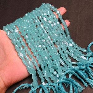 Könnte beinhalten: Eine Sammlung hellblauer Edelsteinperlen, oval geformt mit facettierten Kanten, aufgefädelt. Die Perlen werden in einer Hand gehalten, mit passenden blauen Quasten unten. Die Perlen sind etwa 1,3 cm lang.