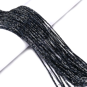 Puede incluir: Cadenas de pequeñas cuentas oscuras y facetadas, probablemente piedras preciosas, se extienden sobre una superficie blanca. Las cuentas tienen un color profundo y reflectante, posiblemente negro o azul muy oscuro, y capturan la luz, creando un efecto brillante.