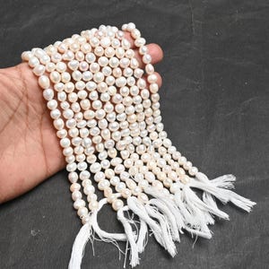 Può includere: Più fili di perle d'acqua dolce bianco crema con una lucentezza lucida. Le perle sono infilate insieme, con nappe bianche alla fine di ogni filo. Le perle sono tenute in una mano su uno sfondo scuro.