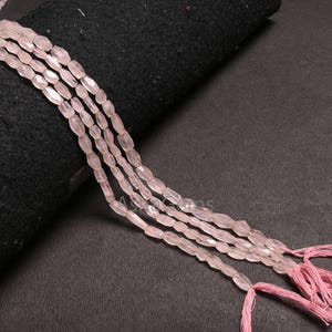 May include: Several strands of pale pink, oval-shaped beads. The beads are strung together, with pink thread tassels at the ends. The beads are displayed on a black surface.