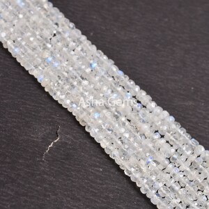 May include: A strand of white moonstone beads, each bead is faceted and has a slightly iridescent sheen. The beads are arranged in a line on a dark gray surface.