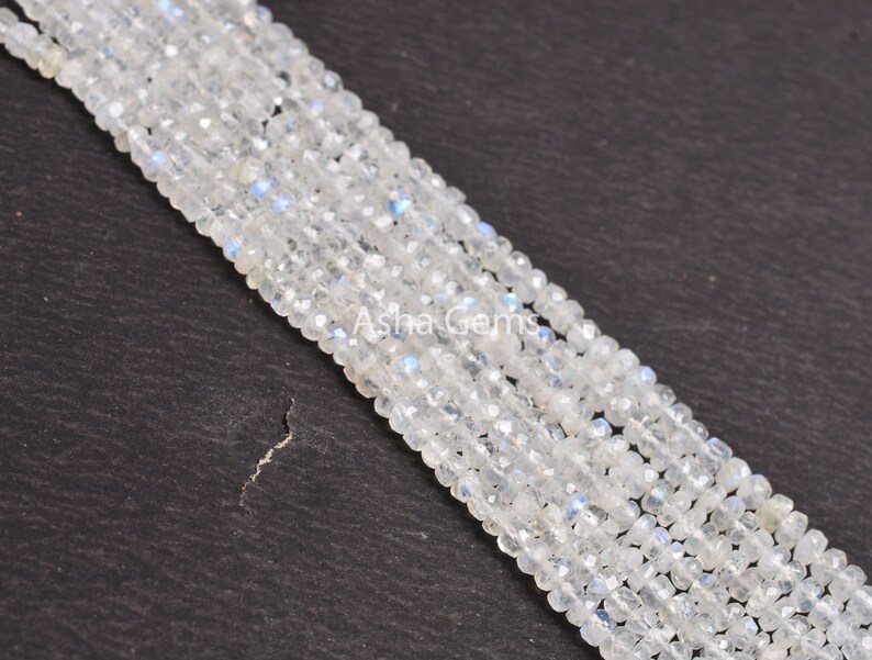 May include: A strand of white moonstone beads, each bead is faceted and has a slightly iridescent sheen. The beads are arranged in a line on a dark gray surface.