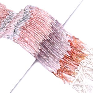 Peut inclure: Gros plan sur plusieurs brins de petites perles rondes dans des tons de rose, pêche, gris et marron. Les perles sont enfilées ensemble, créant une texture fluide. Les extrémités des brins sont finies par des pompons blancs.