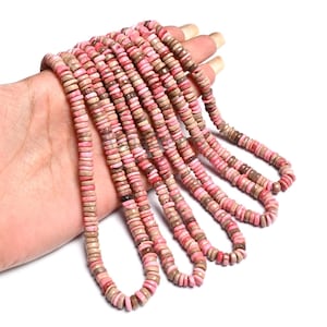 May include: Strands of pink and tan disc-shaped beads. The beads are arranged in multiple strands, creating a layered effect. The beads are held in a hand, showcasing the color variations and texture of the beads.
