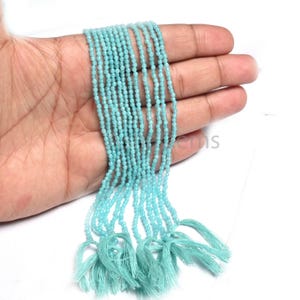 May include: A strand of small, light blue beads held in a hand. The beads are strung together, with tassels at the bottom. The beads are a light, pastel blue color.