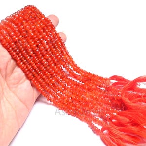 Puede incluir: Varios hilos de cuentas facetadas de piedras preciosas de color rojo anaranjado. Las cuentas están ensartadas juntas, con una borla de hilo naranja a juego al final de cada hilo. Las cuentas se sostienen sobre un fondo blanco.