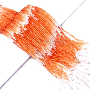 Könnte beinhalten: Stränge aus orangefarbenen und weißen Perlen vor weißem Hintergrund. Die kleinen, gleichmäßig großen Perlen erzeugen eine strukturierte Optik. Die orangefarbenen Perlen dominieren, mit eingestreuten weißen Perlen.