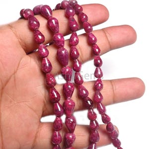 May include: A strand of deep red ruby gemstone beads in a teardrop shape. The beads are strung on a clear thread and are displayed on a hand.