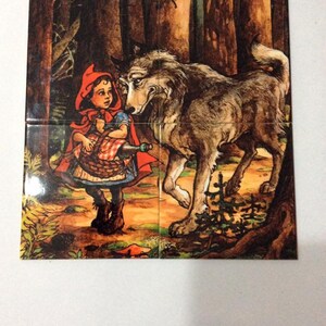 May include: A red hooded girl with a basket and a bottle meets a gray wolf in a dark forest. The girl is wearing a red hooded cloak and a white dress. The wolf is standing on all fours and has a menacing look on its face.
