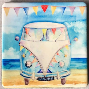 Op de afbeelding: Een blauwe en witte vintage campervan met kleurrijke vlaggen aan de voorkant en een kentekenplaat met de tekst "HAPPY CAMPER". De campervan staat geparkeerd op een strand met blauw water en een zanderige kust.