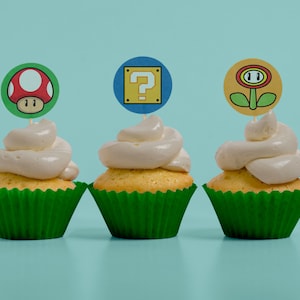 May include: Three cupcakes with green liners and white frosting topped with different video game themed toppers. The toppers are a red mushroom, a yellow question mark block, and a yellow flower.