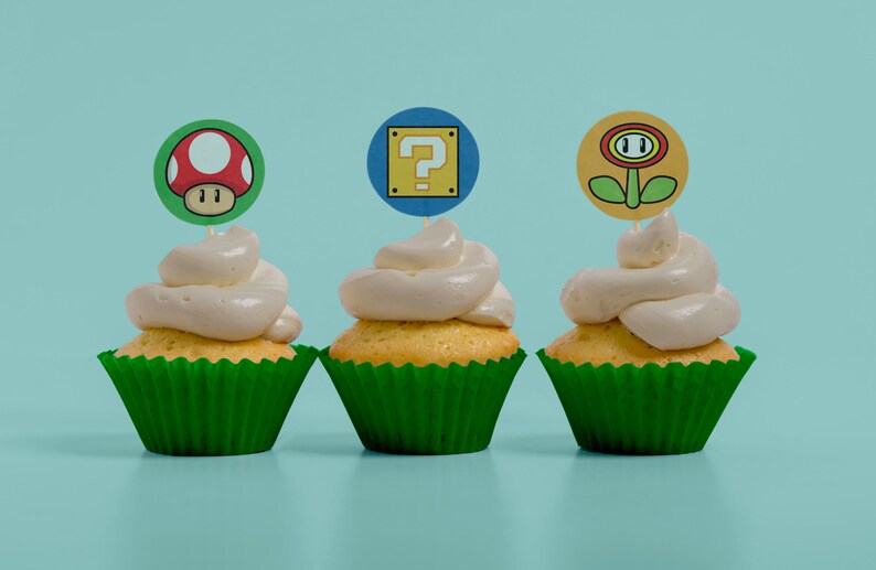 May include: Three cupcakes with green liners and white frosting topped with different video game themed toppers. The toppers are a red mushroom, a yellow question mark block, and a yellow flower.