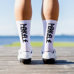 May include: White athletic socks with the word "PERKELE" printed in black. The socks are worn with white athletic shoes. The image is taken from a low angle, showing the feet and lower legs of a person standing on a wooden dock.