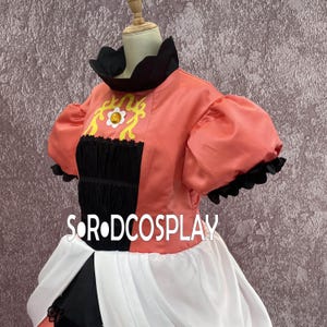 May include: A coral-colored cosplay costume with a black and white skirt. The top features a black collar and sleeves with a ruffled edge. The bodice has a black panel with yellow and white floral embroidery. The text "SOR DCOSPLAY" is visible.