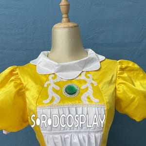 May include: A yellow cosplay dress with white accents, featuring a white collar and puffed sleeves. The dress has a green jewel and white decorative elements on the bodice. The text "SORODCOSPLAY" is visible on the front.