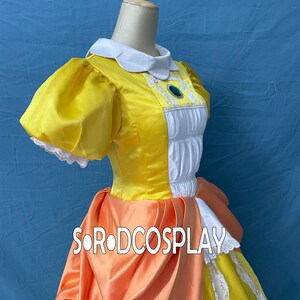 May include: A yellow cosplay dress with a white collar and puff sleeves. The bodice has white panels with decorative trim and a green jewel. An orange sash and skirt overlay add detail. The text "SORODCOSPLAY" is visible.